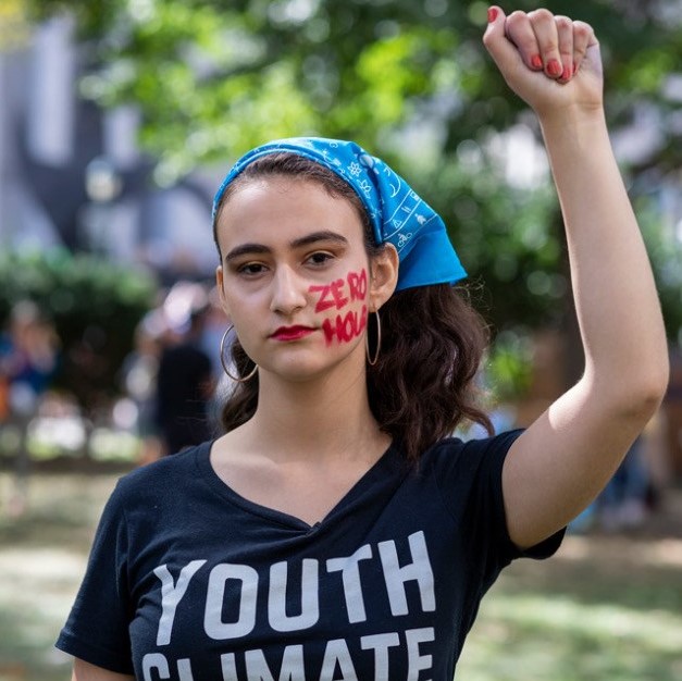 Jamie Margolin stands with fist in the air wearing light blue bandana on head with the words "Zero Hour" written in red lipstick on her left cheek.